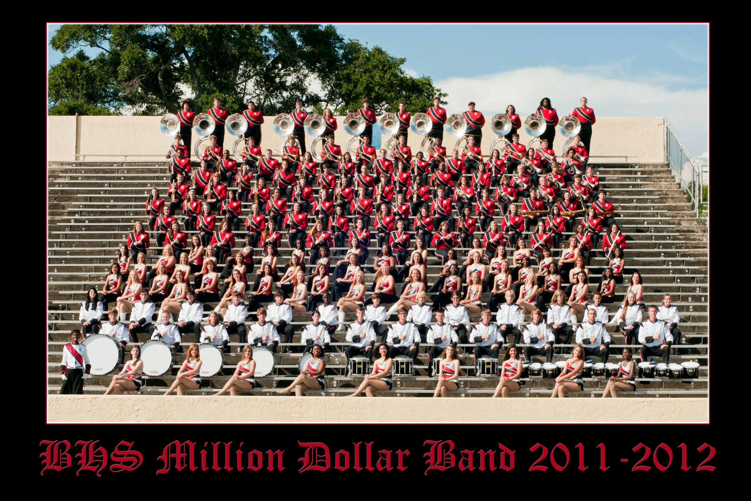 Large marching band performing on outdoor steps with trees in background, dressed in red, black, and white uniforms, with drums and brass instruments.