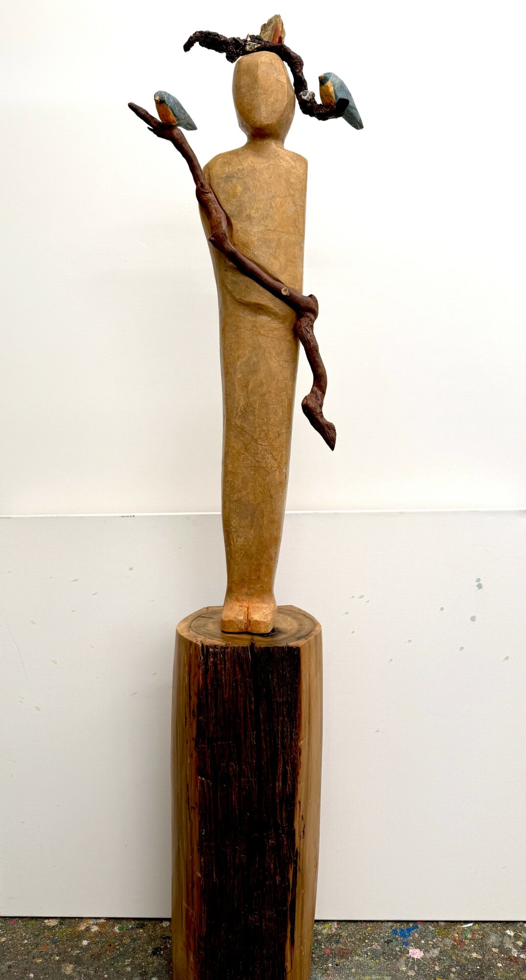 Wooden sculpture of a figure with a branch, head with two small objects, standing on a wooden base, against a white background.