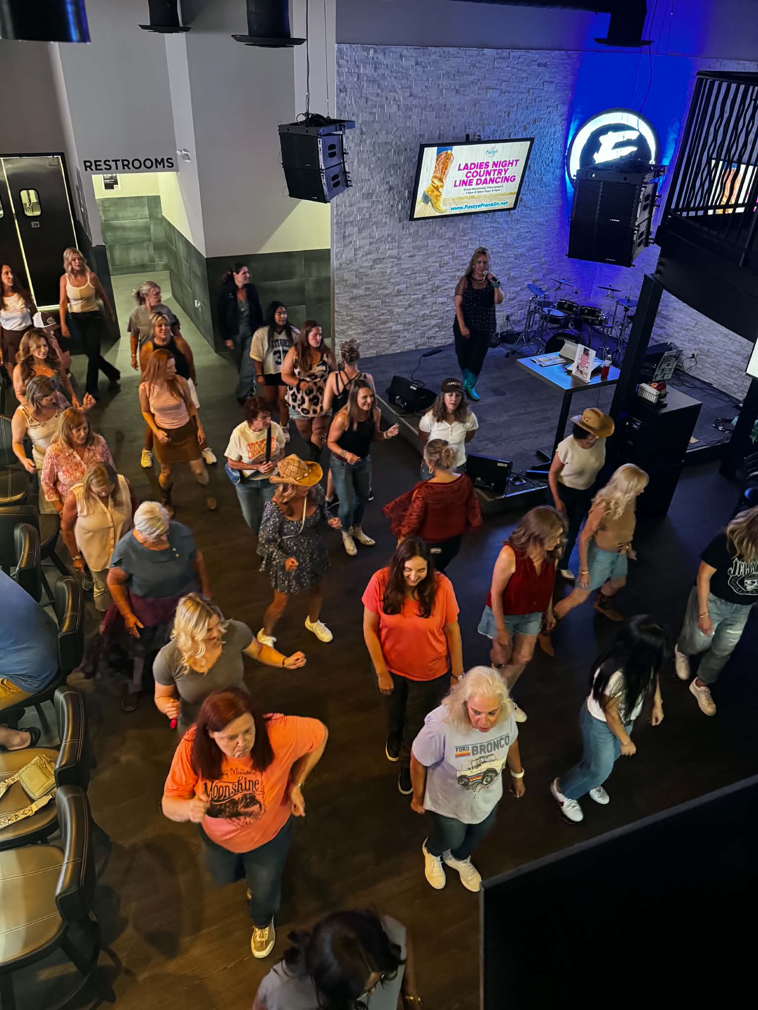 People gathered in a room with a stage, some standing and others walking, with a screen and a sign in the background.