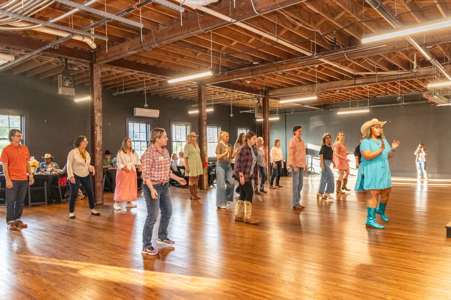 People line up in a spacious indoor dance hall with wooden floors and exposed ceiling beams, some wearing cowboy hats.
