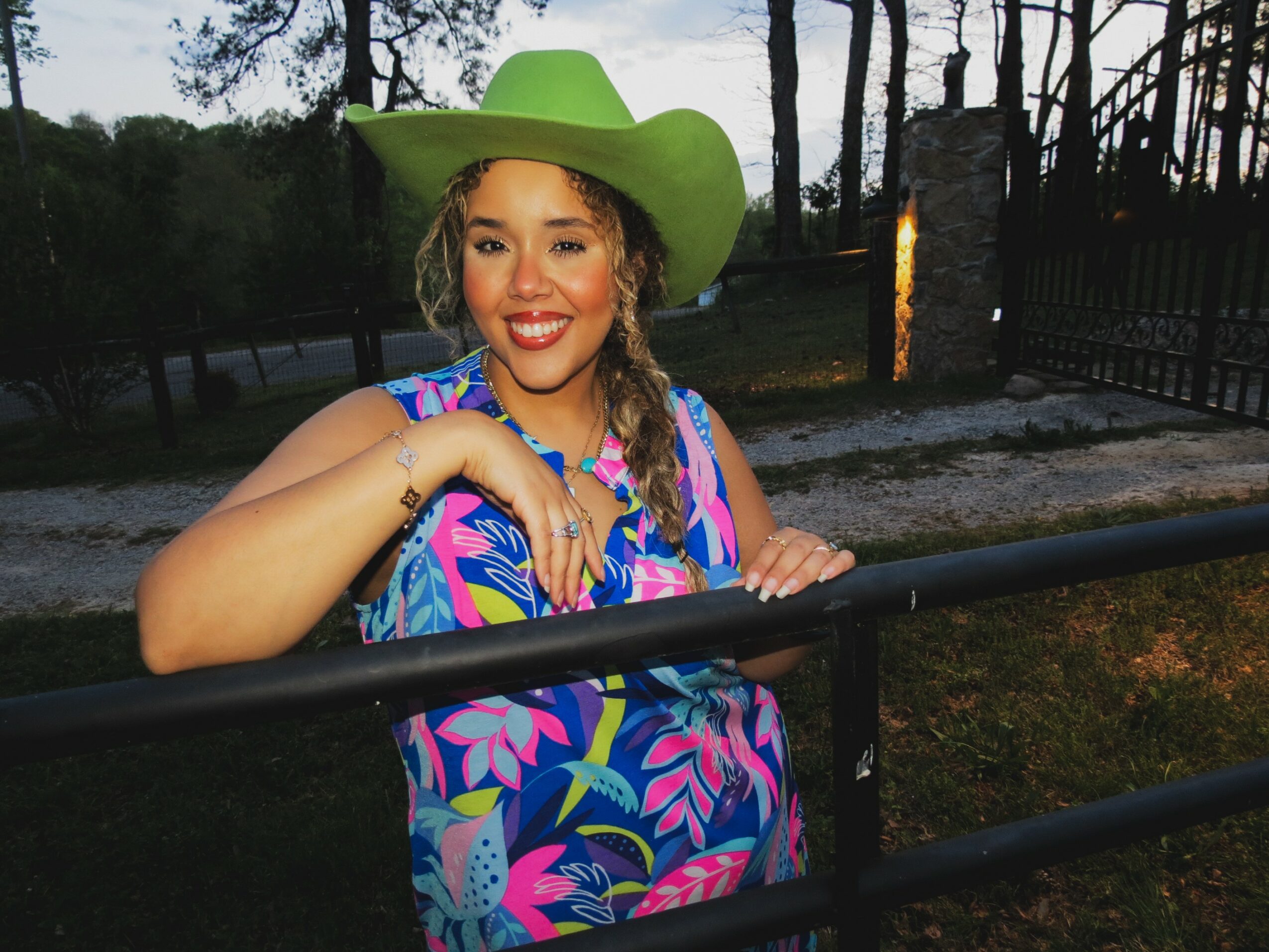Young woman wearing a large green hat and colorful dress, smiling outdoors with trees and a fence in background.