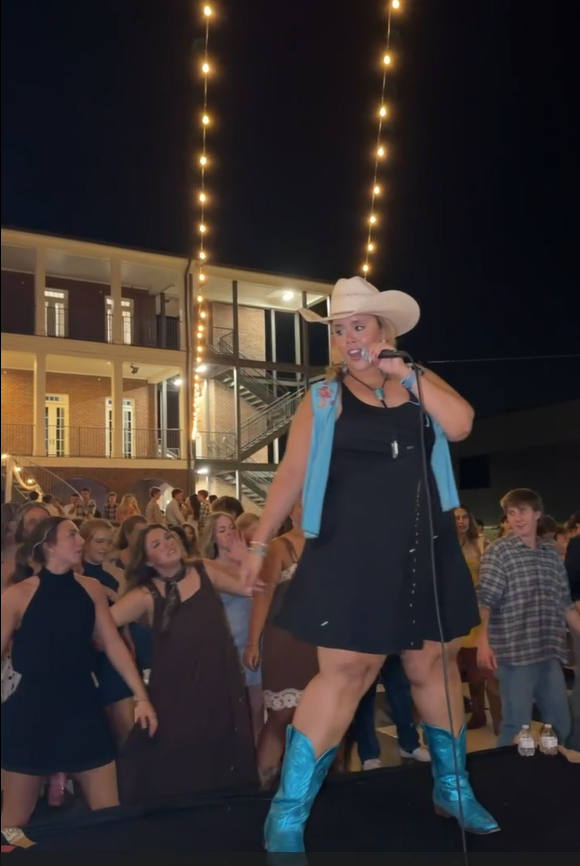 Person in cowboy hat and blue boots on stage at night with crowd and string lights behind.