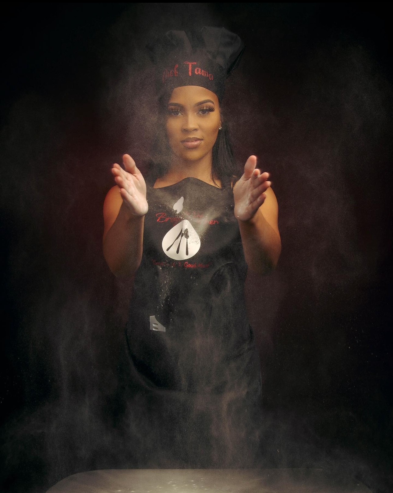 Young woman in graduation cap and gown, holding a rolled diploma, surrounded by smoke or fog, looking at the camera.