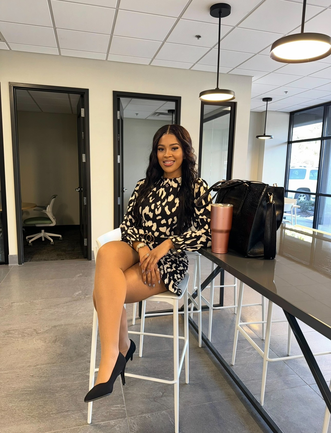 Woman sitting on a high stool in a modern room, smiling, with a black bag and pink tumbler on the table.