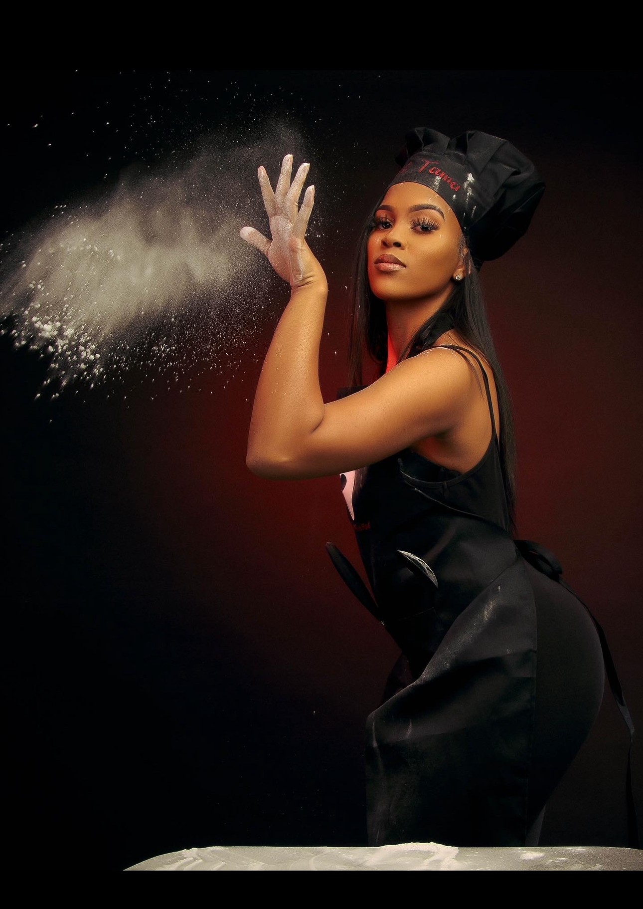 Woman in black chef outfit tossing flour in the air against a dark background, with a confident expression.