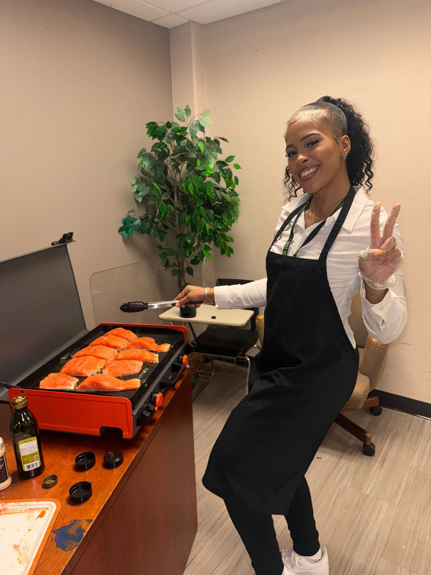 Woman in apron making sushi, smiling, holding a peace sign, standing next to a table with sushi and cooking equipment.