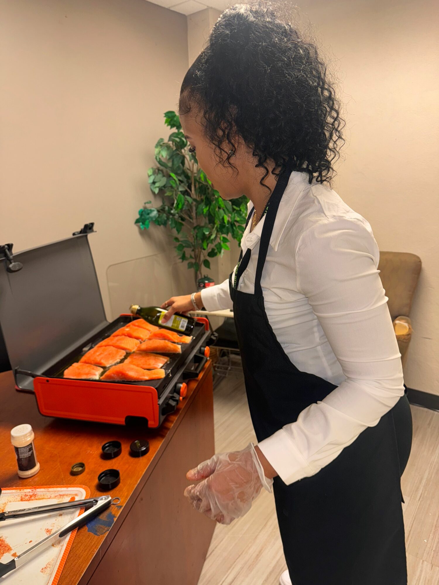 Person with curly hair wearing a white shirt, black vest, and gloves, preparing food on a table with a tray of salmon fillets.