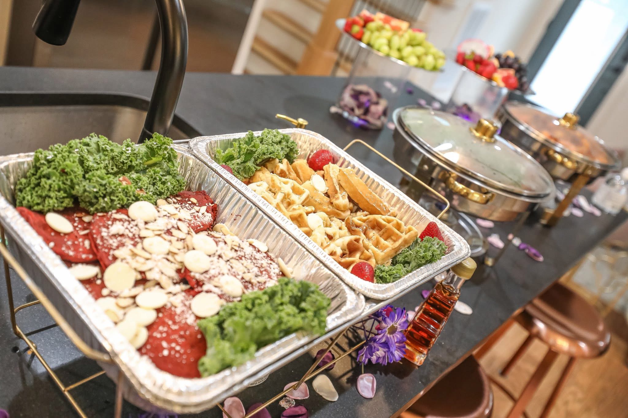 Buffet table with trays of sliced tomatoes, cheese, and greens, along with waffles and toppings, in a dining area.