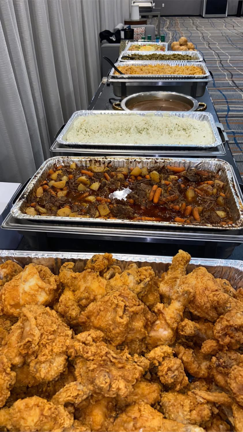 Line of food trays with fried chicken in foreground, various dishes behind, on a table outdoors, viewed from the front.