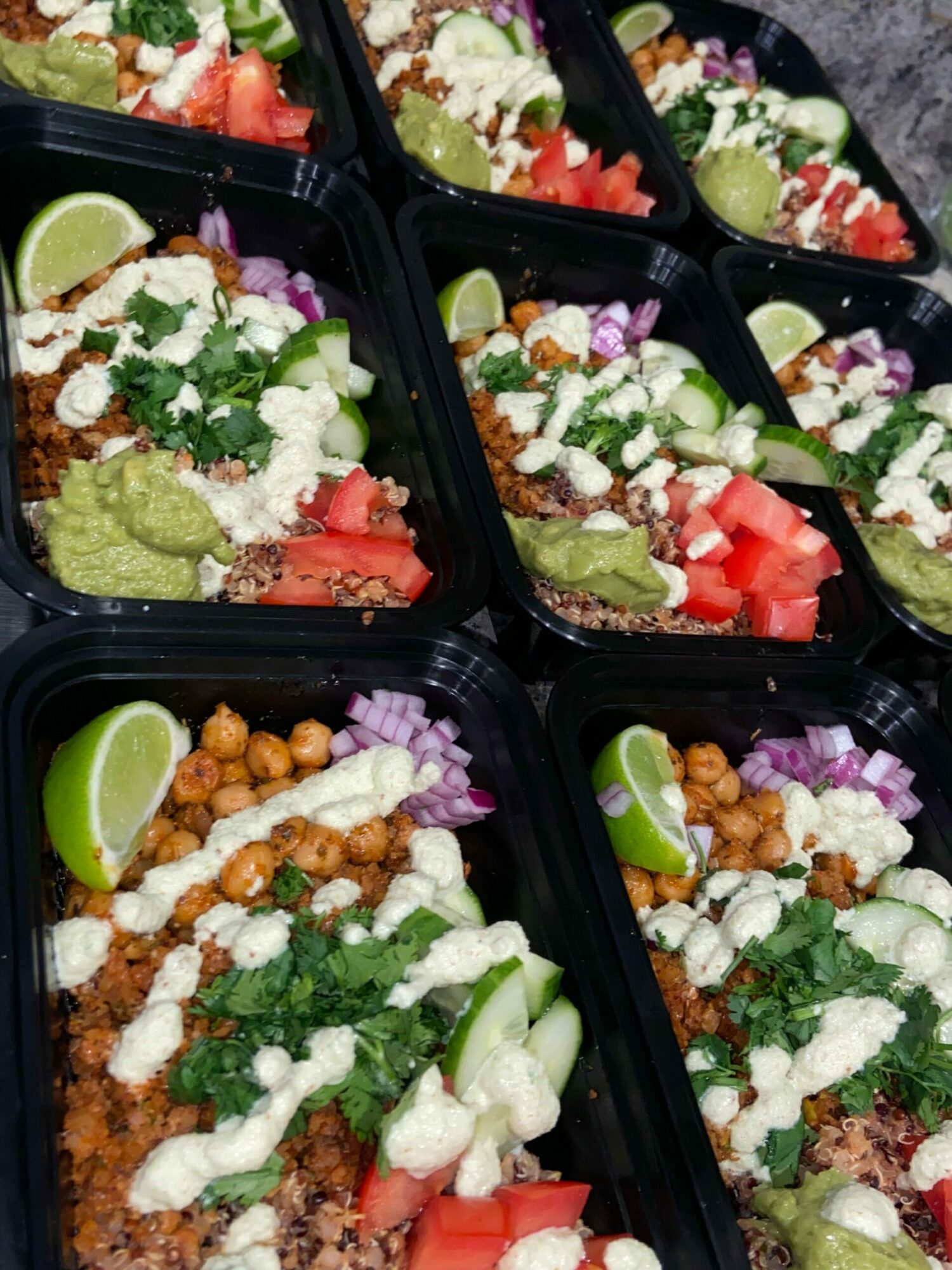 Multiple black containers filled with tacos topped with lettuce, cheese, tomatoes, onions, and lime wedges, arranged on a surface.
