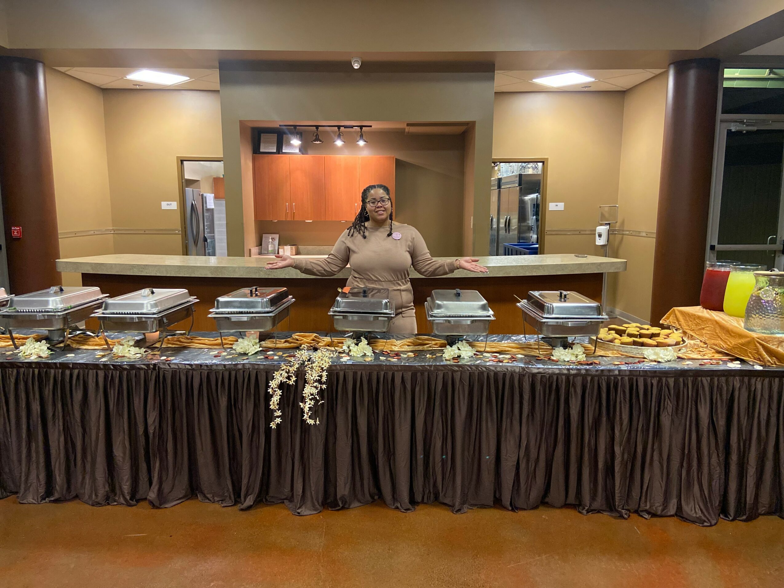 A woman stands behind a buffet table with food trays in a hotel or conference room, smiling and gesturing with her hands.