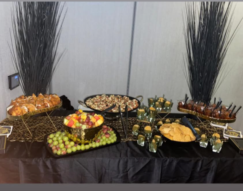 Dessert table with various sweets, fruits, and snacks, decorated with tall black plants on each side, against a plain background.