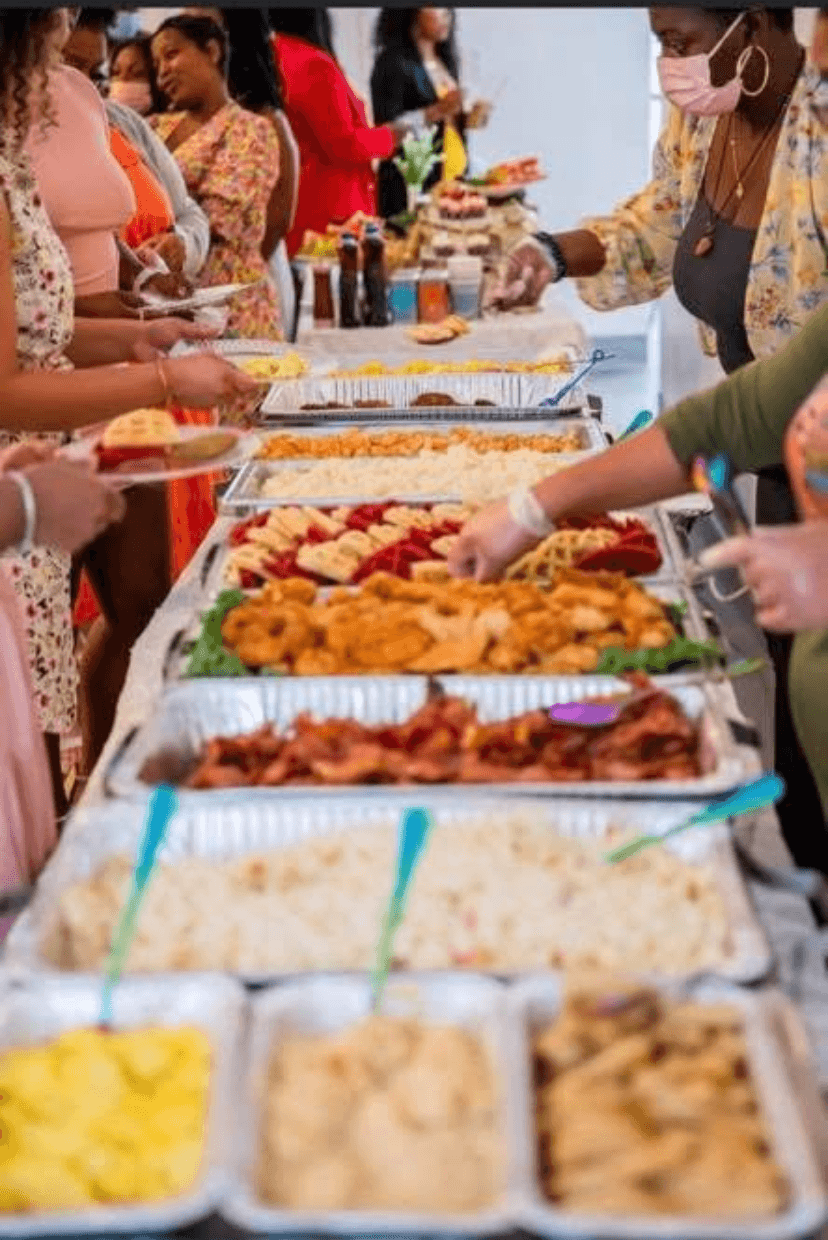 People gather around a buffet with various food trays, serving and selecting dishes at a social event.