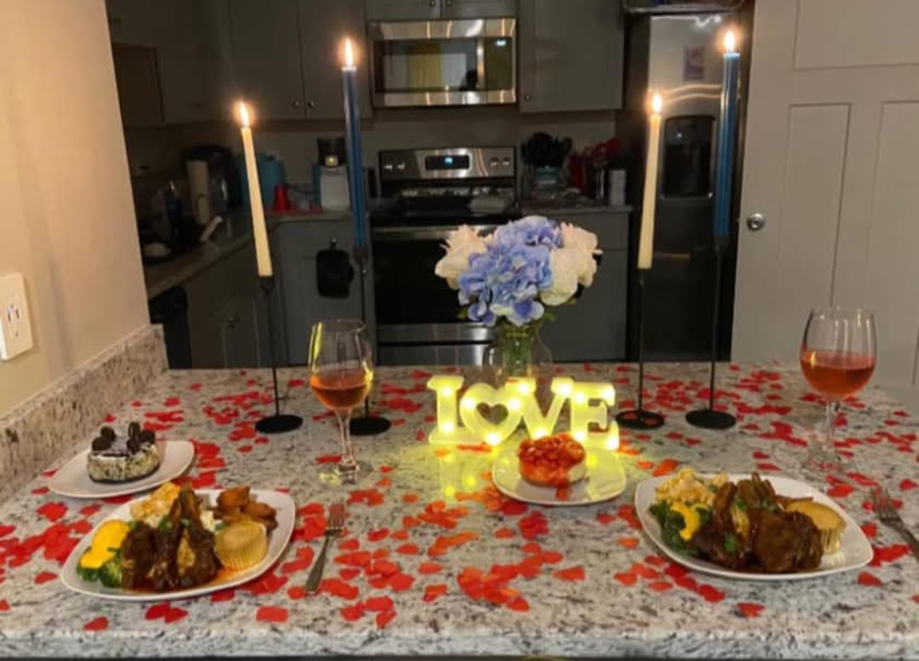 Valentine's Day dinner setup with candles, flowers, wine glasses, and plates of food on a kitchen counter, decorated with rose petals.