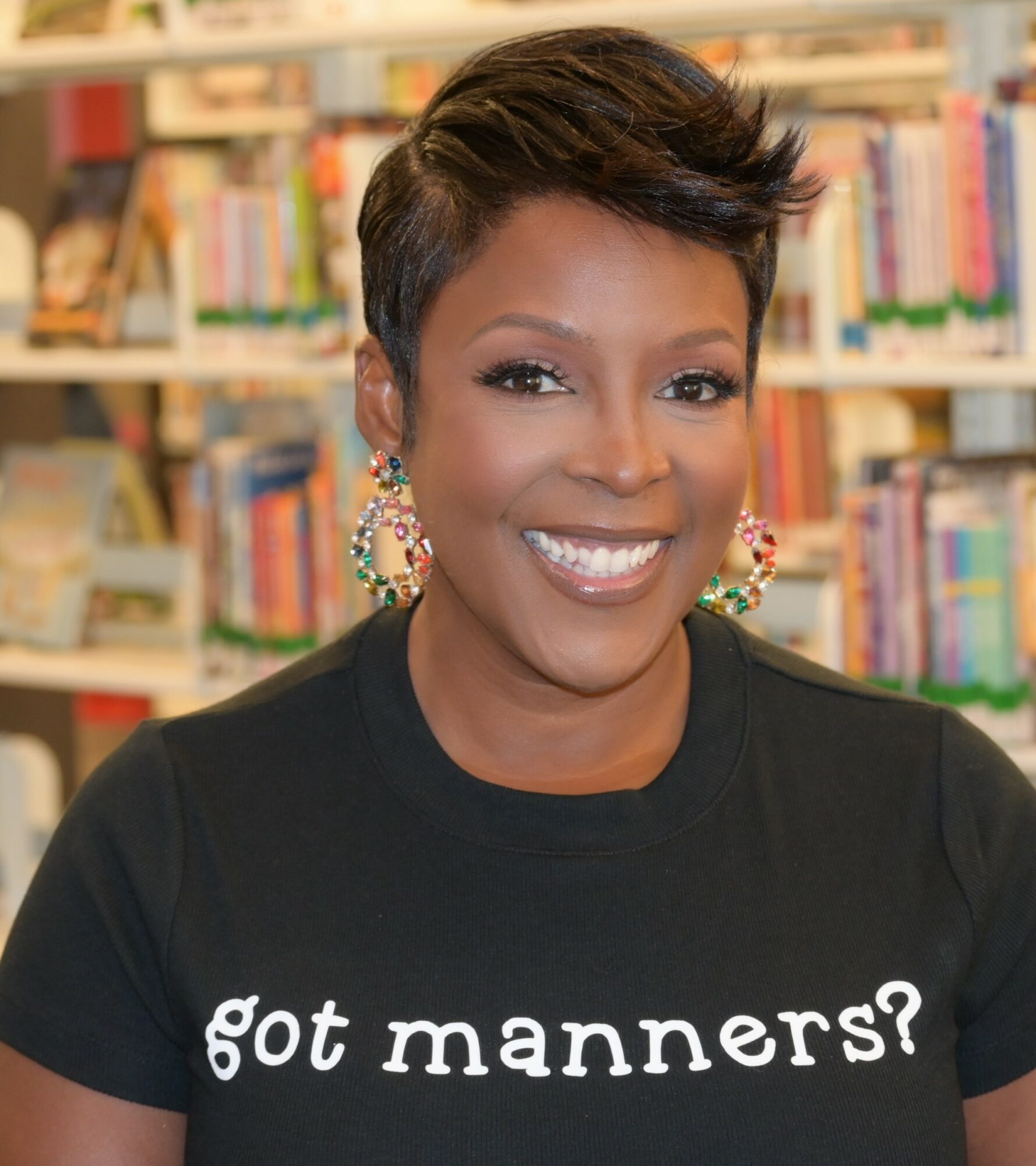 Smiling woman with short hair and earrings in a library, wearing a black shirt with 'got manners?' text.