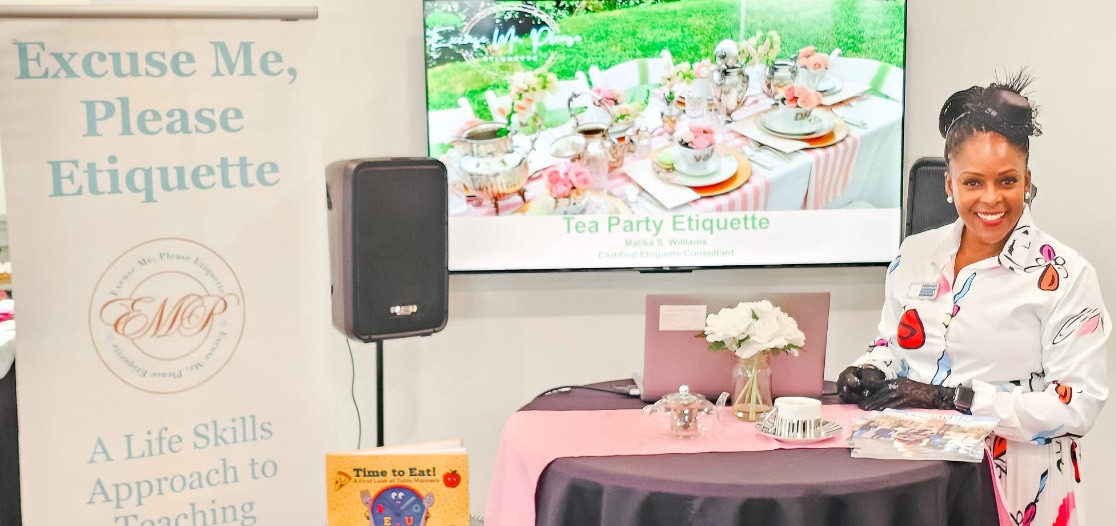Woman in uniform smiling at a table with a pink cloth, flowers, and a microphone, in front of a screen displaying a tea party scene.
