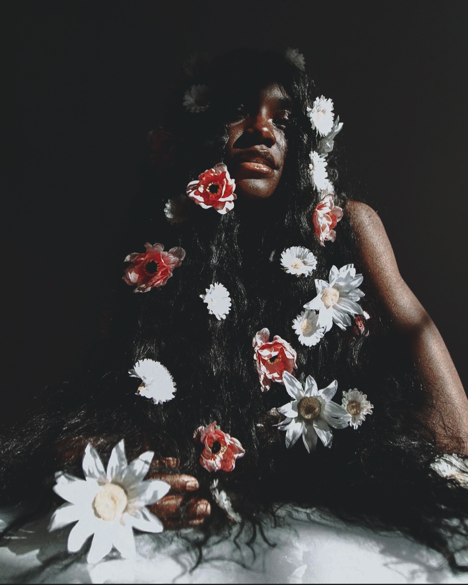 Woman with long hair decorated with flowers, dark background, serious expression, flowers on hair and around her.