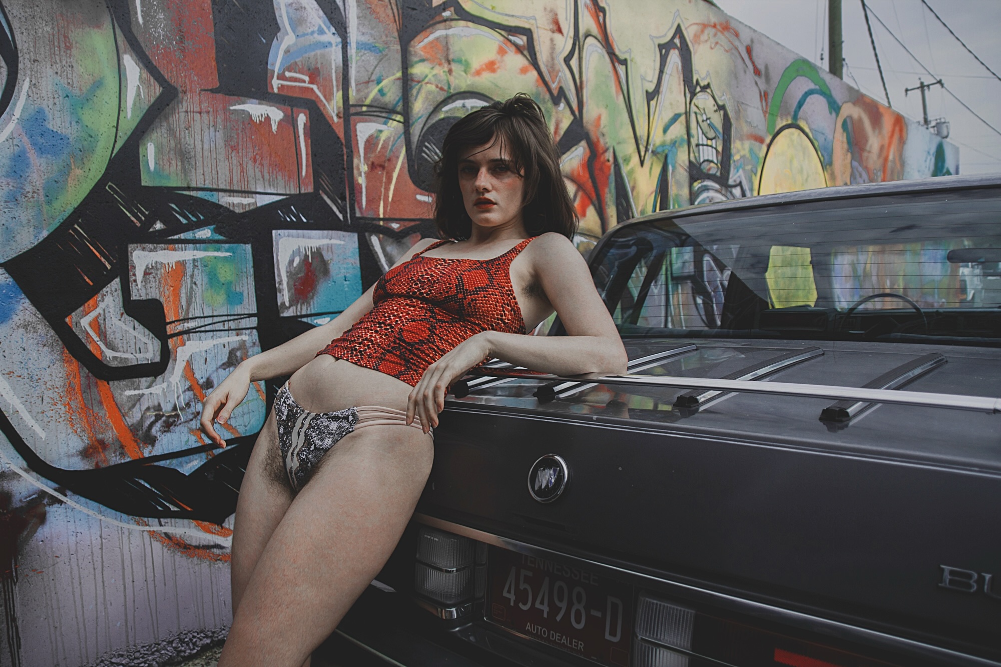 Woman with dark hair and sunglasses leaning on a black car in front of a colorful graffiti wall, wearing a red top and shorts.