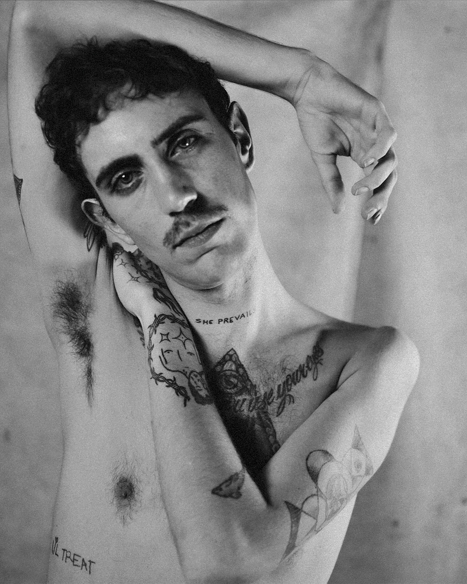 Black and white portrait of a shirtless person with tattoos, resting their arm on their head, looking at the camera.