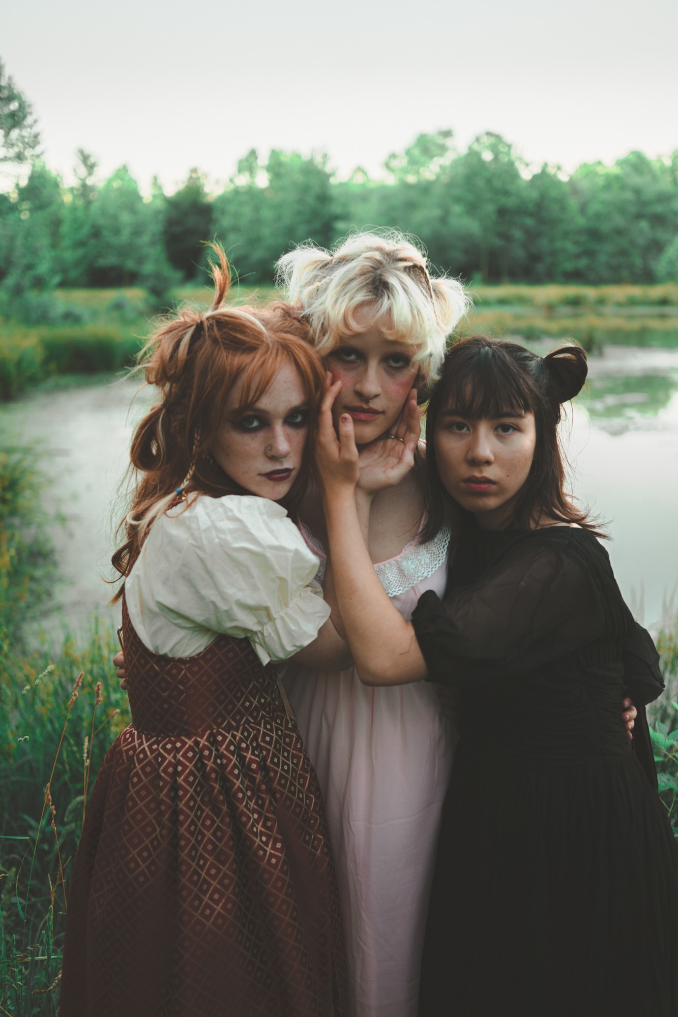 Three women with different hair colors and styles standing outdoors near a body of water, with trees in the background.