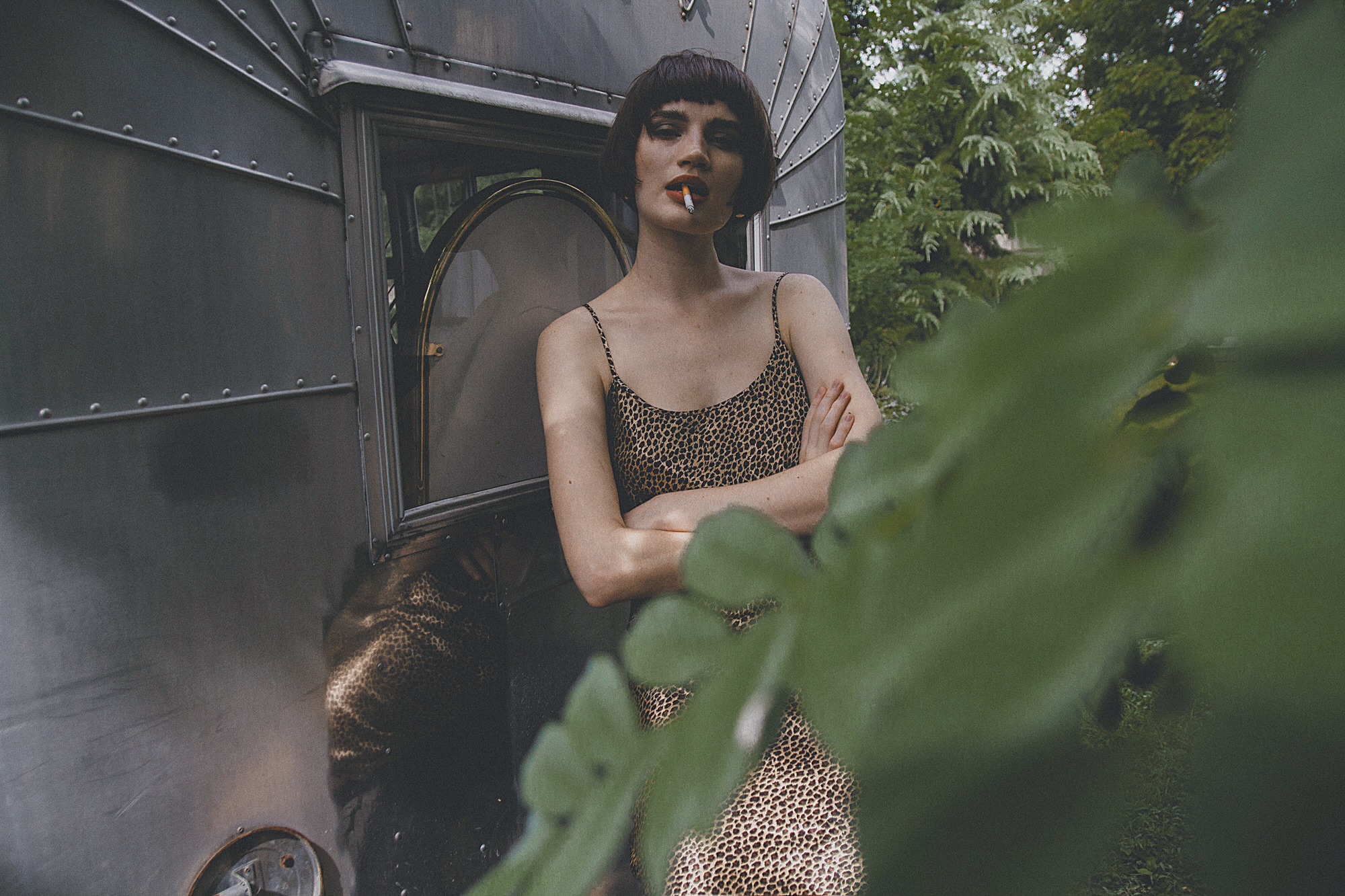 Woman with short dark hair in a leopard print dress standing outside a metallic structure, partially obscured by large green leaves.
