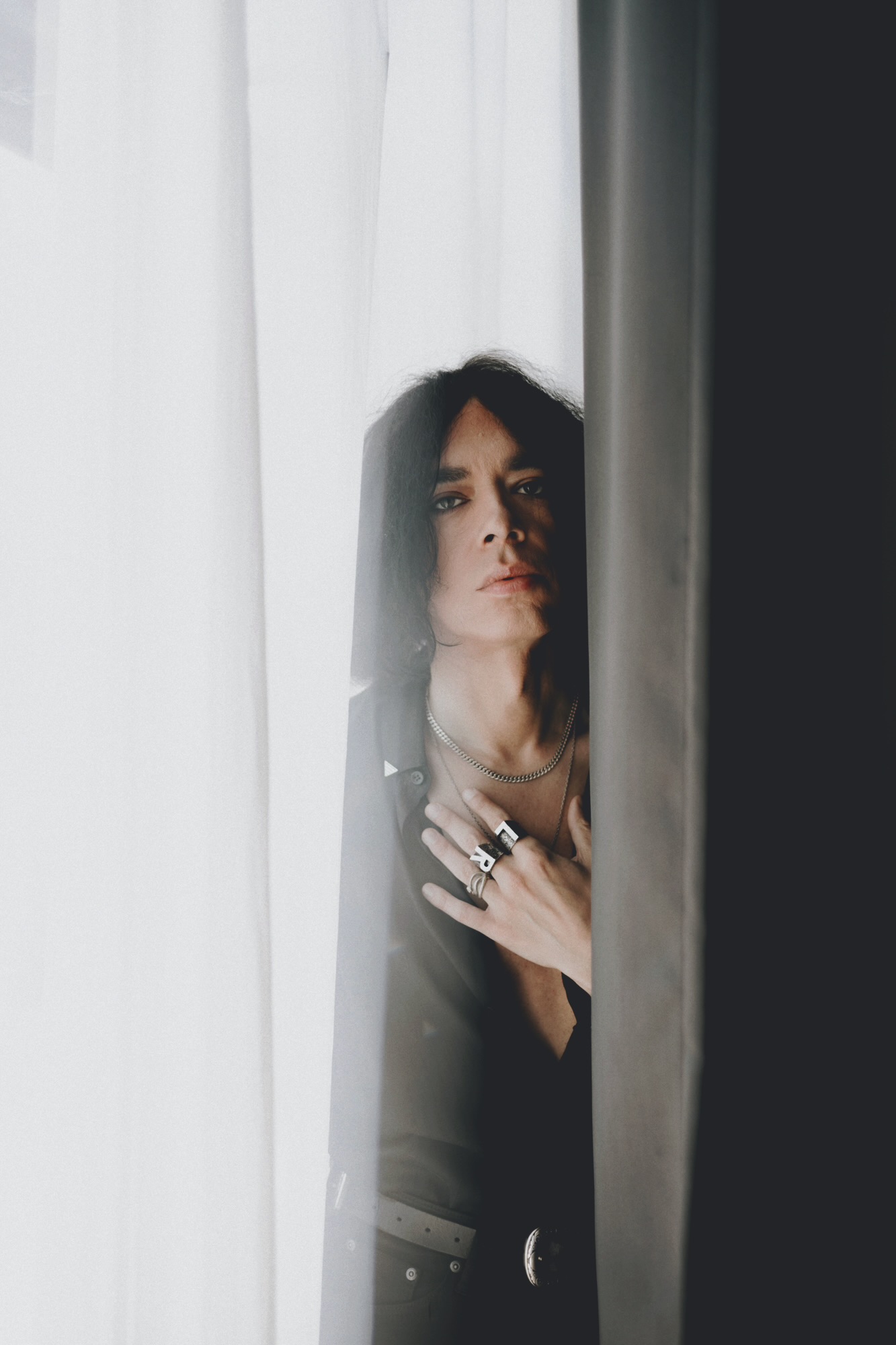 Woman peeking through curtains, partially obscured, with dark hair and rings on her fingers, looking at the camera.