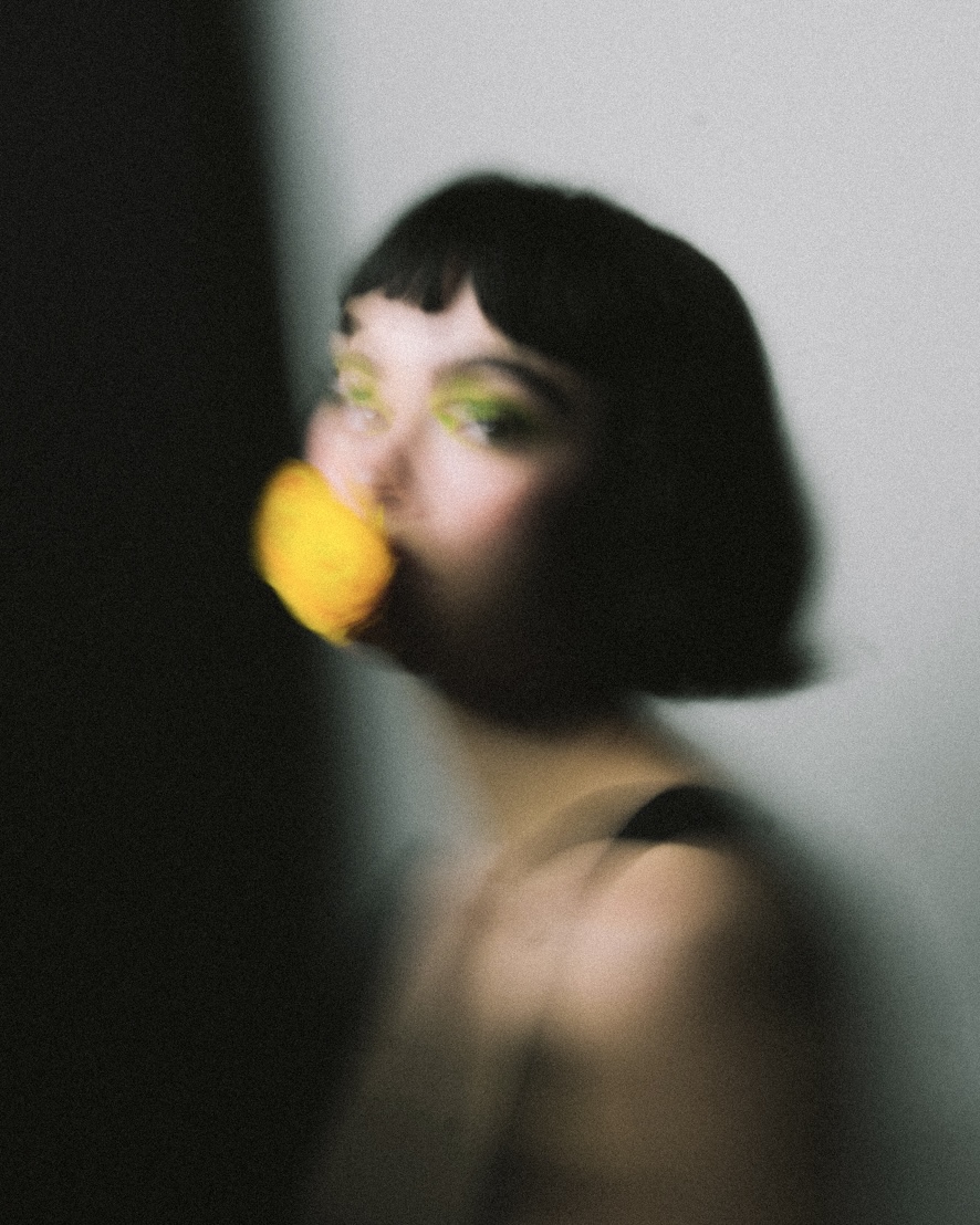Blurred portrait of a woman with short black hair, yellow eyeshadow, and a yellow object near her face.