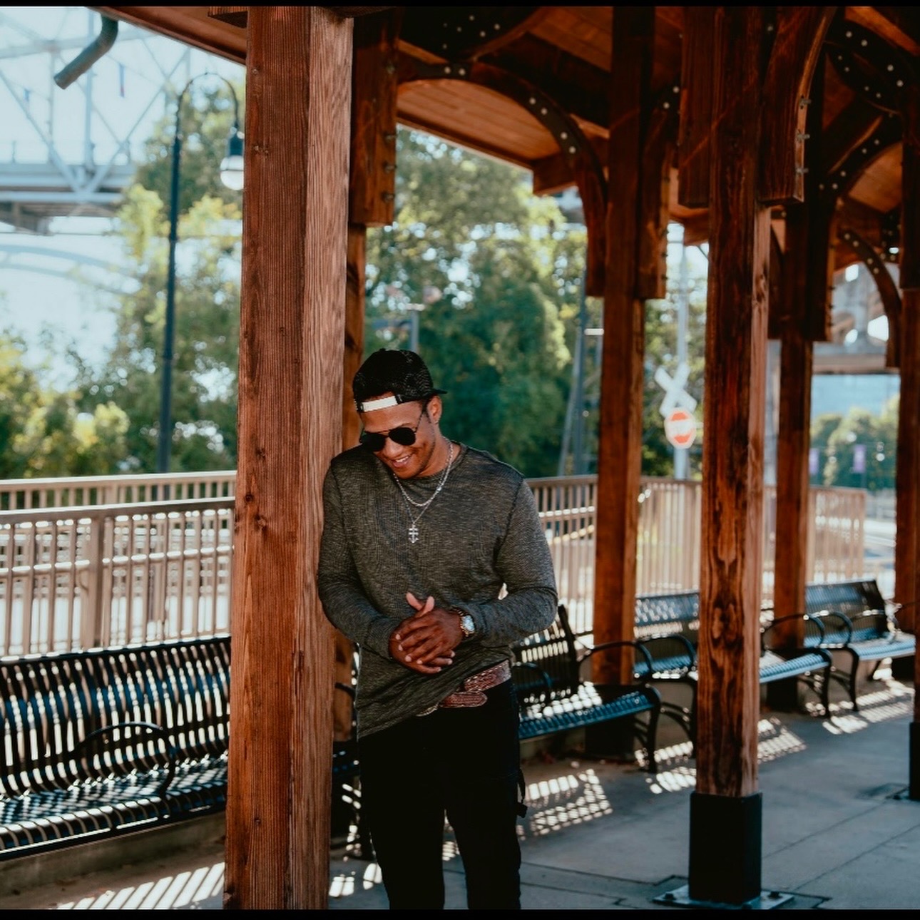 Person standing outdoors under a wooden structure, wearing sunglasses and a dark long-sleeve shirt, with trees and benches nearby.