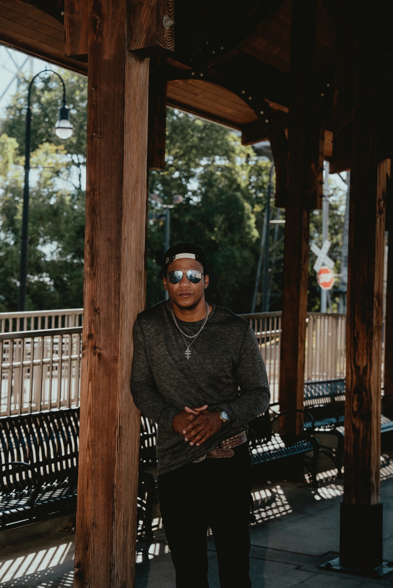 Person wearing sunglasses and dark clothing standing under a wooden structure outdoors, with trees and a fence in the background.
